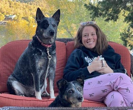 Meredith sitting on a deck with her two blue heeler dogs, Laguna and Sailor, holding a cup of coffee