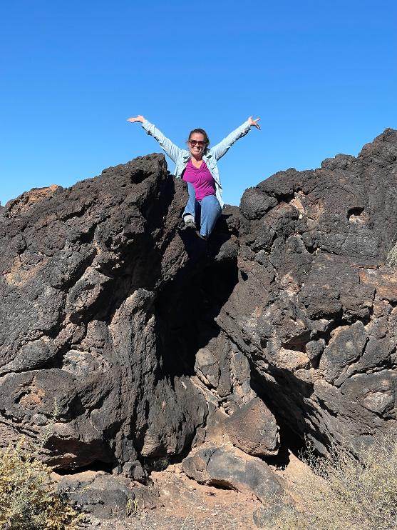 Meredith standing on a black lava rock formation in New Mexico with both arms raised under a clear blue sky